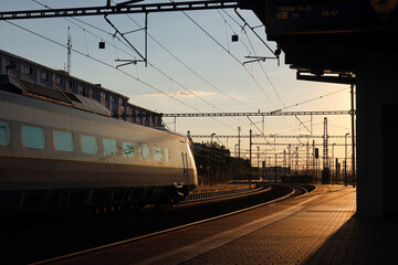 A high-speed train passes through Prague Liben station at sunset, reflecting warm evening light. Overhead power lines frame the sky, and long shadows stretch across the calm platform.
