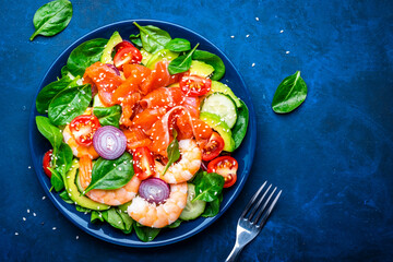Healthy food salmon salad for keto diet with shrimp, avocado, spinach, cucumber, tomato, cashew, sesame seeds. Blue background, top view