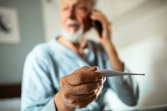 Elderly man checking thermometer while calling doctor at home