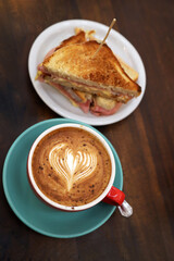 Large cup of coffee on a bar table with a toasted ham and cheese sandwich. Red bowl with heart-shaped latte art. Hot drink. Coffee with milk. Cappuccino.