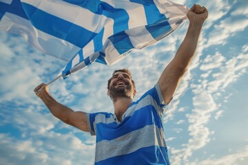 Man stands in joy, holding a Greece flag against a blue sky dotted with white clouds. His arms raised high, he wears a blue and white striped shirt matching the flag colors.