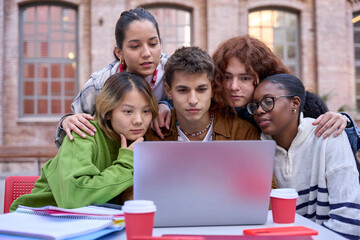 Front view of group of students working together using and looking concentrated at laptop outdoor. Studying for exams and sharing knowledge sitting at table on university campus. Education technology