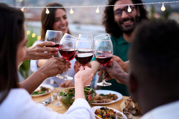 Focus on red wine glasses of unfocused young people toasting with alcoholic drinks gathered celebrating summer party on rooftop. Happy group friends enjoying smiling together sitting at meal table
