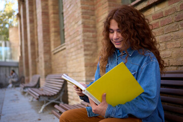 Smiling young Caucasian with red hair non-binary student sitting on bench outside university building looking at notes and holding notebook. Generation z nice people campus lifestyle. Copy space