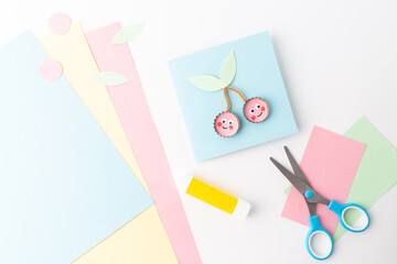 Flat lay of craft materials on pastel background: pink, yellow, blue paper, cherry cutout, green leaves, scissors, bottle cap. Creative DIY project setup,