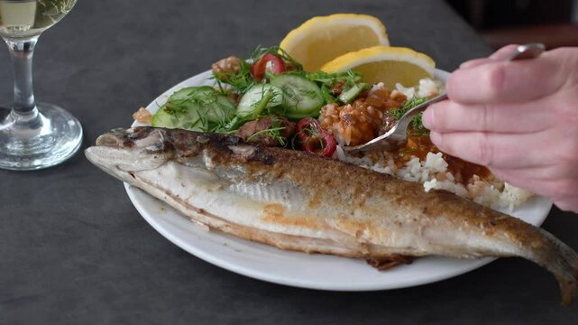 unrecognizable man eating fried sea bass with cutlery served with rice, fresh salad and lemon wedges, glass of white wine on table, concept of varied and tasty food in all inclusive hotel,