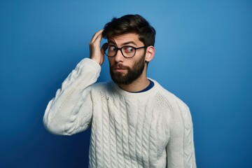 Fototapeta premium Young handsome man with beard, glasses wears casual sweater over blue background. Confused, uncertain, he wonders about question, thinking with hand on his head. Pensive, thoughtful expression.