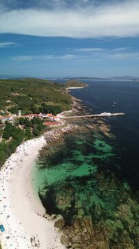 Pristine Area dos Cans beach with turquoise waters, surrounded by rocks, leads to Ons village. A catamaran rests by the dock, while scenic hills and cliffs stretch to Melide Beach under sunny skies.