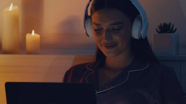 Woman gamer in bed playing multiplayer game on laptop at night, wearing headphones