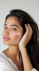Fototapeta premium South Asian woman applies white cream on her skin in a beauty studio with a gray background, focusing on hand holding the cream. The facial application showcases intimate atmosphere and relaxed state.