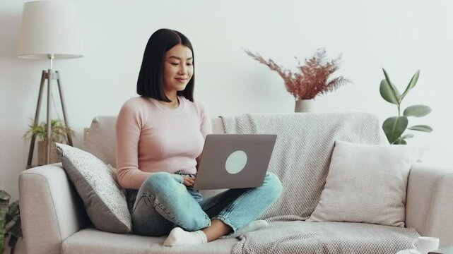 Young asian woman networking on laptop at home, freelancing online at home, sitting on couch, free space