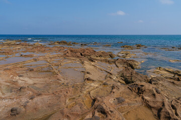 A rocky coastal landscape featuring shallow tide pools under a clear blue sky. The serene ocean meets the rugged shore, creating a peaceful natural scene