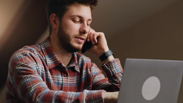 Online consultation. Young male manager typing on laptop and talking with client via cellphone, working remotely at home, slow motion