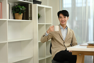 Fototapeta premium Positive young entrepreneur in suit drinking hot coffee at modern office