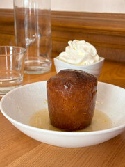 A plate of dessert called Baba au Rhum in a plate on table with a bowl of whipped- cream and clear glass in background