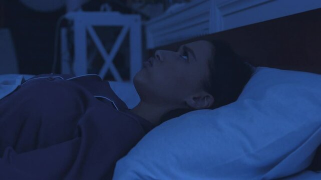 Young irritated woman trying to sleep, annoyed by bad noisy neighbors, covering ears with pillow in bed late at night