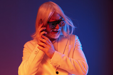 Stylish older man in a white suit, wearing sunglasses and talking on a smartphone against a vibrant blue red gradient background, exuding confidence and modernity