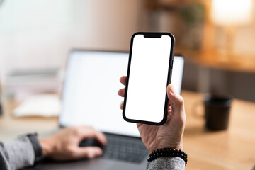 Hand holding texting using mobile on desk at home office, cell phone blank white screen mockup. Background empty space for advertise.
