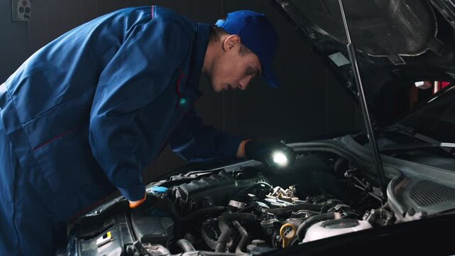 Professional auto mechanic examining car engine, looking under hood with flashlight, side view