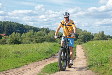 Cyclist in helmet with equipment on electric bicycle rides along path in field in countryside....