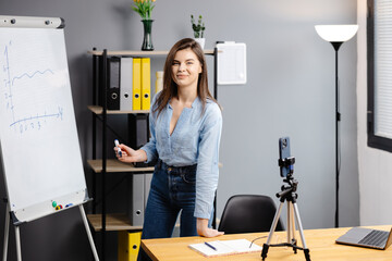 Young woman mentor recording presentation, webinar on smartphone standing on tripod, teacher coach...