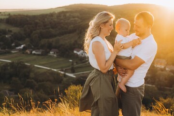 Happy Family Enjoying Sunset in Scenic Countryside Landscape, Smiling Parents Holding Baby in Warm Evening Light