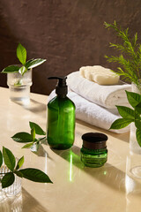 Natural spa theme photo with unlabeled green pump bottle and moisturizer pot placed by side on light yellow spa table top, next to some glass jars contain green plant, bath towel and soap