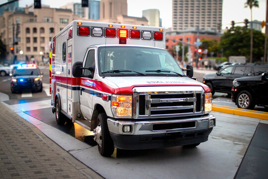 An ambulance with lights activated and a police car behind it  in an urban environment