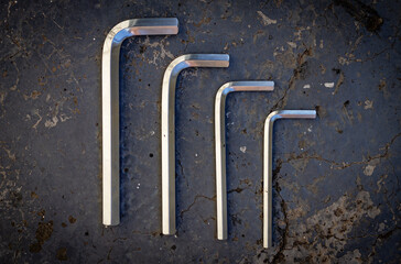 A set of steel hex keys arranged from large to small for assembly work on a dark gray cracked concrete surface. Color horizontal photo from top to bottom with a plain background.