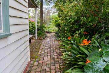 A narrow brick pathway beside a wooden house or country cottage leads into a lush, quaint, and well-maintained garden. Cozy and peaceful atmosphere in a quiet country home.