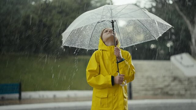 Happy teenage girl in a yellow jacket holds an umbrella and watches water drops flow down it during heavy rain while walking in the park