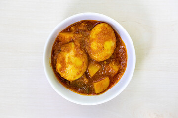 Spicy egg curry or Dim Vuna, cooked with potato, in a white ceramic bowl, placed on a light wooden surface. Top view of flavorful boiled dim bhuna, a Bengali recipe often enjoyed with rice or bread.
