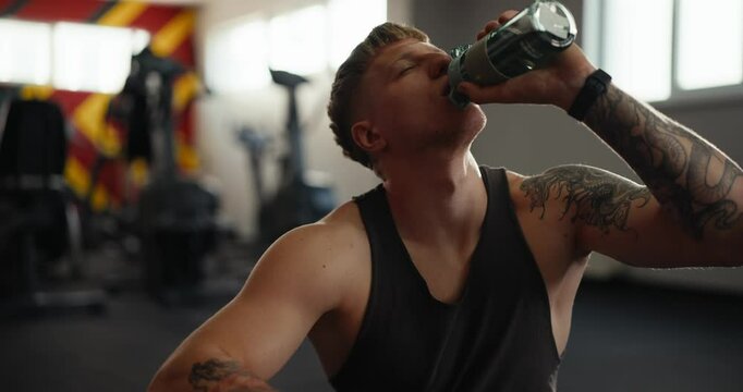 Up Close confident guy blond athlete tattoo in a gray T-shirt drinks water from a sports bottle to maintain water balance in the body in the gym