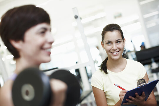 Coach, woman and clipboard with dumbbell for workout at gym, notes and test for progress at gym. Personal trainer, people and writing on checklist with stats, fitness and exercise at wellness club - Powered by Adobe