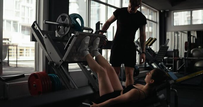 A brunette athlete girl in a sports uniform does leg press exercises with weights that her trainer set up for her during a workout in the fitness room