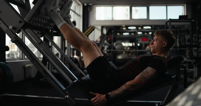 Side view of a confident guy athlete in a black sports uniform with tattoos and in gray sneakers performs a bench press during his leg workout in the fitness room