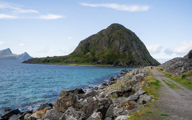 A beautiful coastal landscape showcasing a rocky shoreline with a prominent green hill rising by the sea. The clear blue water gently laps against the rocks, and the background features distant mount