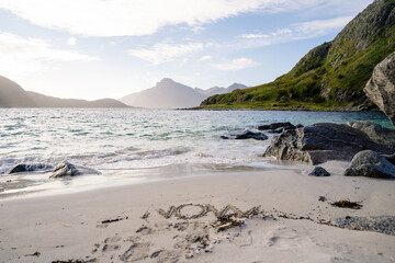 A beautiful coastal landscape showcasing a rocky shoreline with a prominent green hill rising by the sea. The clear blue water gently laps against the rocks, and the background features distant mount