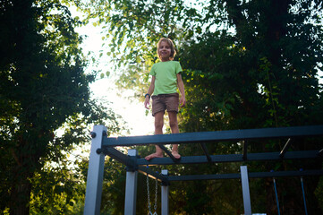 Fototapeta premium A child joyfully stands on a playground structure surrounded by lush greenery in the late afternoon sunlight