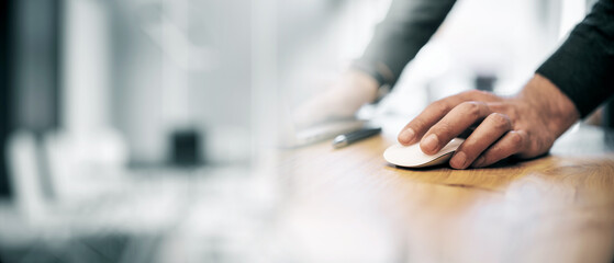 Closeup of a man's hand using a computer mouse on a wooden desk in an office setting.
