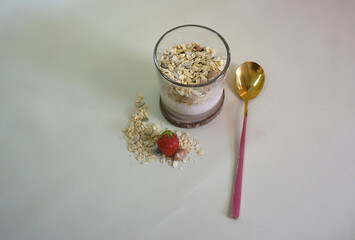 Top View Of Glass With Yoghurt, Muesli, And Oats Spoon On White Table.