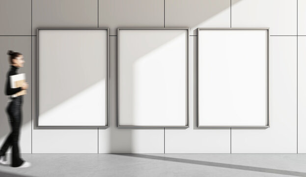 Three vertical blank posters on a wall with a woman walking by, holding a notepad, in a modern building.