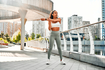 Portrait of an attractive athletic woman with a fit figure in a white top who has stopped after jogging and exercising outdoors, resting and listening to music in headphones. Sports lifestyle.