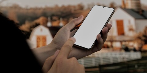 Caucasian White male using smartphone with a blank white screen. Farm, sunset, barn apps mockup. Vertical phone orientation