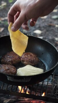 Vertical footage of male hands adding cheese on meat burger cutlet on pan at outdoors. Person preparing meal dinner. Making tasty hamburger dish on bonfire in autumn forest