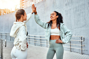 Sporty women give each other a high five while standing outdoors before a workout and run, against a grey urban wall.