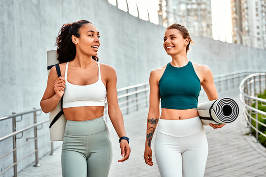 Attractive modern sports women walk holding mats for fitness training, yoga and joyfully chatting. Sincere friendship and sports lifestyle.