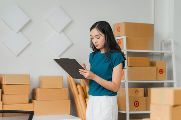 Focused Businesswoman in Home Office:  An Asian entrepreneur stands in her home office, meticulously reviewing a clipboard amidst a backdrop of cardboard boxes, symbolizing the hustle and dedication o