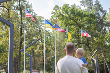 Man holding little child against Ukrainian and American flags. Rear view, selective focus.