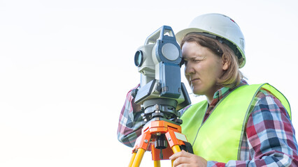 A girl in a white helmet near a surveying instrument looks into the telescope with her right eye...